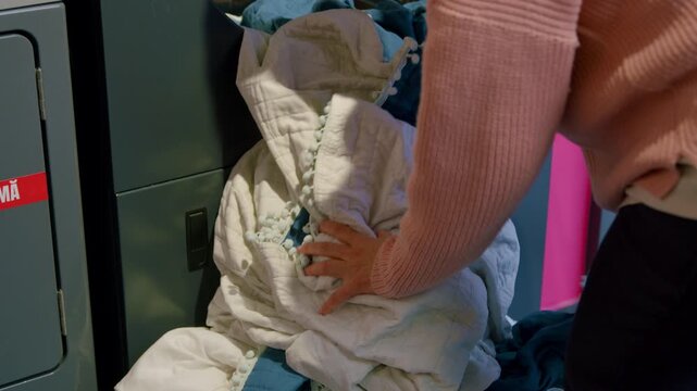 Woman in pink sweater loads large white comforter into washing machine at self-service laundromat, preparing for laundry in a bright, modern setting