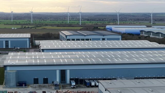 Biggleswade, United Kingdom - 24 January 2026: Aerial view of warehouses reflecting the overcast sky, juxtaposed against distant wind turbines on the horizon.