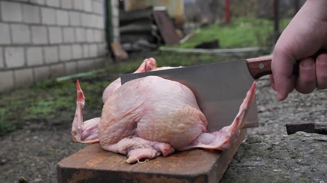 A butcher cuts up a chicken carcass with a kitchen cleaver on a cutting board for preparation.