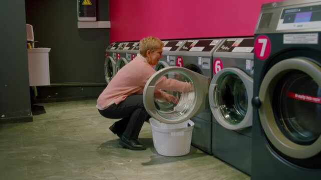 Individual with short hair in pink sweater loads laundry into washing machine at self-service laundromat, showcasing process of washing clothes in modern facility