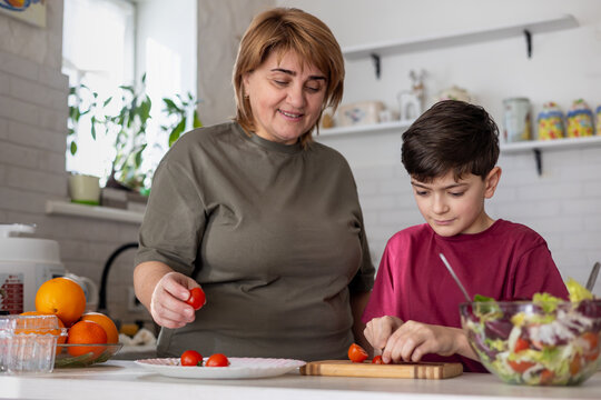Senior woman cooking healthy meal with her grandson at home. family meal preparation, learning and intergenerational bonding. Authentic domestic lifestyle scene focused on healthy food, care, together
