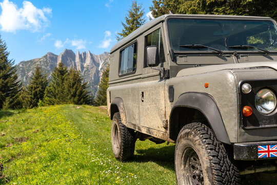 Pararul Recce, Romania - 07 June 2025: View of a rugged, mud-splattered off-road vehicle parked on a vibrant green meadow, with towering, jagged mountain peaks piercing the clear blue sky.
