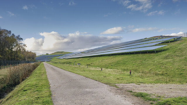 Wijster, Netherlands - 27 October 2024: View of solar panels glinting atop a verdant landfill hill, contrasting with the textured grey pathway and a sky streaked with clouds.