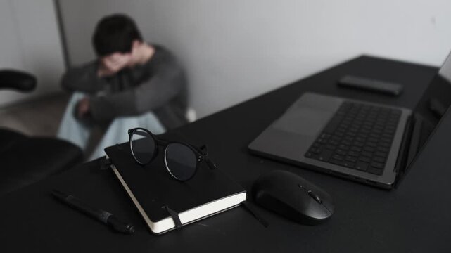 Depressed man with head down sitting on floor in despair in home office next to open laptop and glasses, illustrating mental fatigue. Realistic depiction of freelance burnout.
