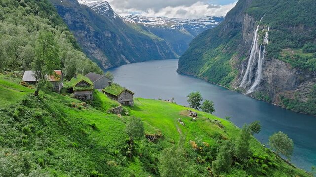 Drone view of Skagefl&aring; village perched above Geirangerfjord with wooden structures and grass roofs. Expansive Norwegian fjord landscape surrounds the remote settlement.