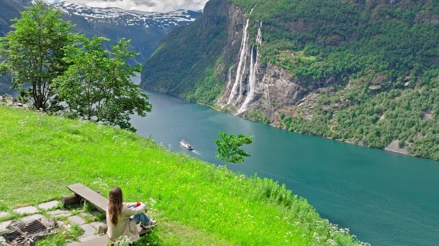 Drone view of a female traveler sitting on a hillside bench overlooking the Seven Sisters Waterfall and Geirangerfjord in Norway. Calm summer daylight emphasizes Norwegian fjord scale, emerald slopes.