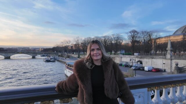 A woman in a fur coat takes candid selfies on a bridge in Paris The Seine river flows below with boats passing by and the city skyline is visible in the background under a blue sky during daytime.