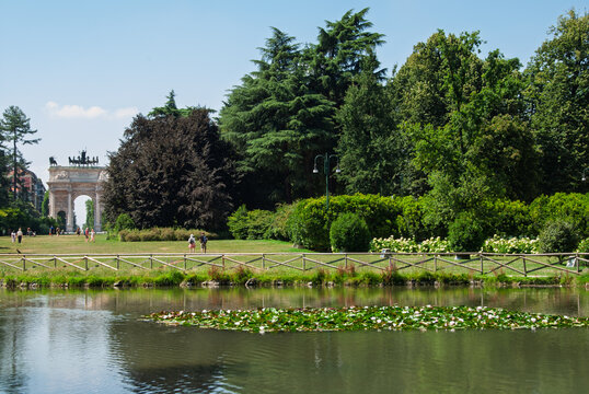 Peaceful view of Parco Sempione in Milan with a reflective pond, lush greenery and the Arco della Pace in the background on a sunny summer day.