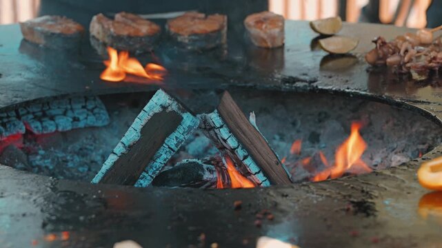 Close-up of delicious fish and seafood cooking on a hot metal plancha around a wood-burning fire pit. The warm glow of flames illuminates the scene, creating a rustic and appetizing atmosphere