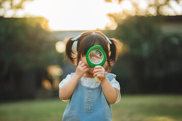 Curious Asian toddler girl looking through a magnifying glass in a sunny garden. Childhood discovery exploration and outdoor learning concept. Little explorer playing in the park during golden hour