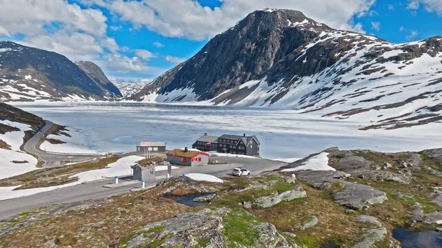 Aerial view approaching a Norwegian mountain hotel complex with a visible national flag near a frozen alpine lake. Clear daylight reveals snow patches, access roads, and rugged highland terrain.
