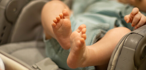 Close-up of a baby's feet resting in a car seat. The infant has chubby toes and soft skin, wearing a light blue outfit. The background is softly blurred.
