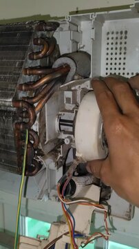Air conditioner service close up showing a hand reaching into an open indoor unit with copper pipes fan motor wiring and dusty components during residential cooling system repair work