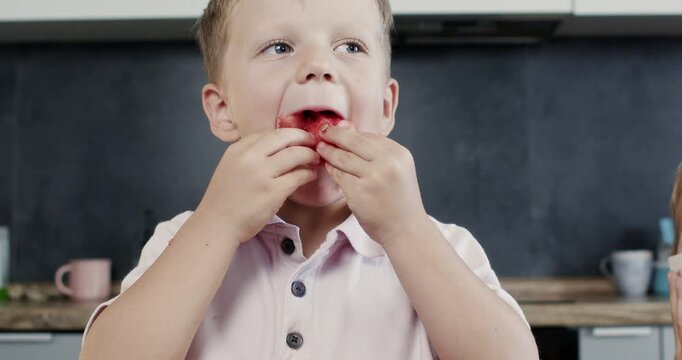 Young boy shoves watermelon chunk into mouth at kitchen table chewing fast. Preschooler child pinches rind with hands searching seeds carefully on lips before next bite