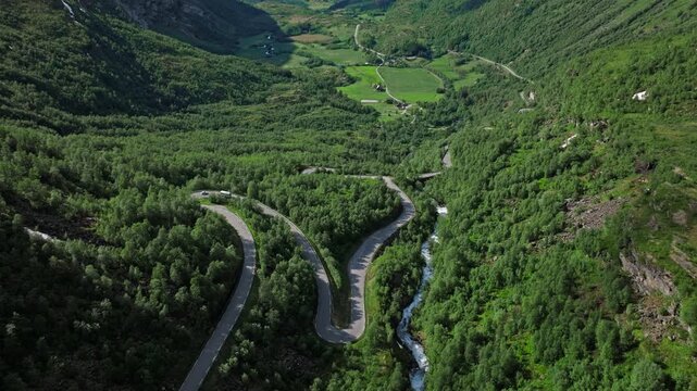 Elevated aerial shot following sharp hairpin bends of a Norwegian mountain road cutting through dense forest in Norway. The scene emphasizes dramatic terrain, flowing streams, and classic Norwegian.