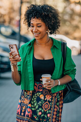 woman with afro hair walking on the street with mobile phone and cup of coffee