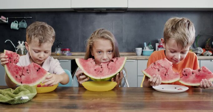 Children sit at kitchen table chewing watermelon wedges from bowls. Boy in orange t-shirt grins biting slice hard while sister chews near younger boy hiding behind rind