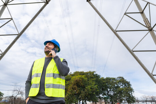 Engineer wearing safety gear talking on smartphone near electricity pylon