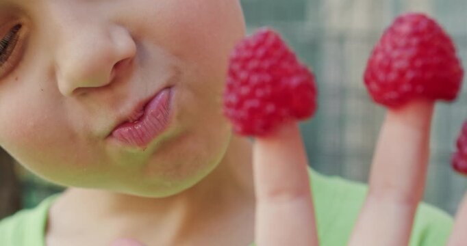 Young boy opens mouth biting raspberry covering fingertip. Child chews berry licking juice while hand withdraws to grab next fruit during fresh snack time