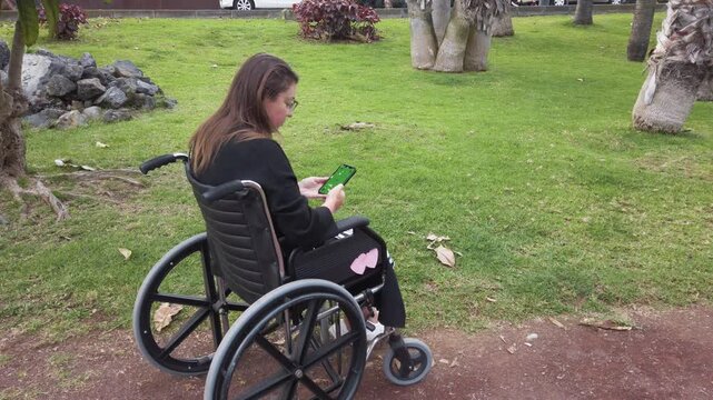 woman in wheelchair using smartphone, student scrolling messages along leafy park path green grass and trees border dirt trail, soft daylight and quiet mood,