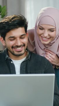 Handsome positive bearded guy sitting on comfortble sofa in living-room while his splendid cheerful wife in headscarf standing behind him and they revisioning together intersting program on laptop