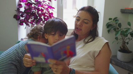 Obraz premium Mother reading picture book with sons in living room, toddler watching and smiling while older child concentrates on pages, warm educational moment