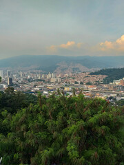 Obraz premium The city of Medellín at the base of green hills in a valley, featuring tall buildings and a busy downtown street grid. Distant mountains mixed with a soft, hazy sky above.