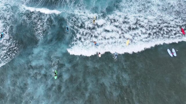 Aerial view of surfers riding waves in the ocean. Multiple unrecognizable people on surfboards navigating through the water, with dynamic wave patterns and foamy surf visible in the background