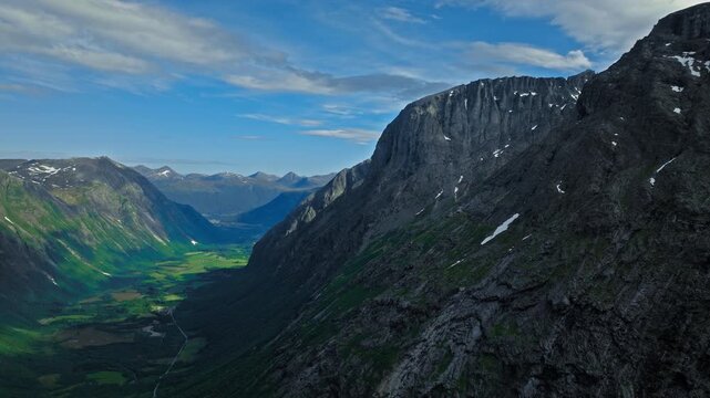 Drone view opens to a wide valley at Trollstigen framed by towering cliffs and green lowlands. Beyond Trollstigen&rsquo;s rock walls, distant hills and layered terrain complete the scene.