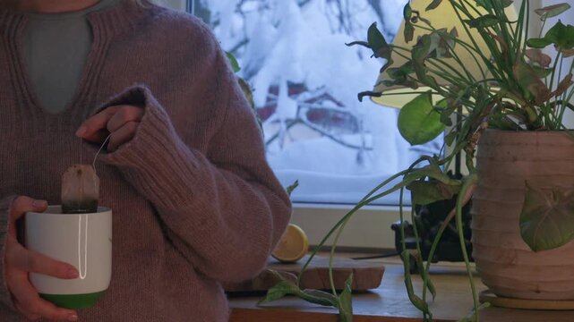 Close-up of a woman holding a cup of tea while standing with her back to the window in a cozy winter interior. Warm light and snowy view outside create a calm atmosphere of comfort, mindfulness and qu
