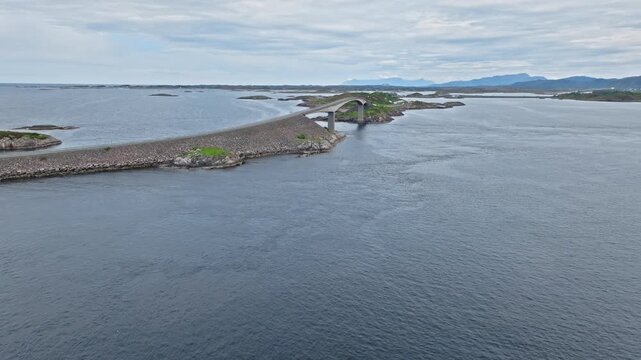 Atlanterhavsveien aerial view of the road causeway and bridge spans crossing between low skerries and open water. Drone view slides over the straight rock berm and bridge approach, revealing the route
