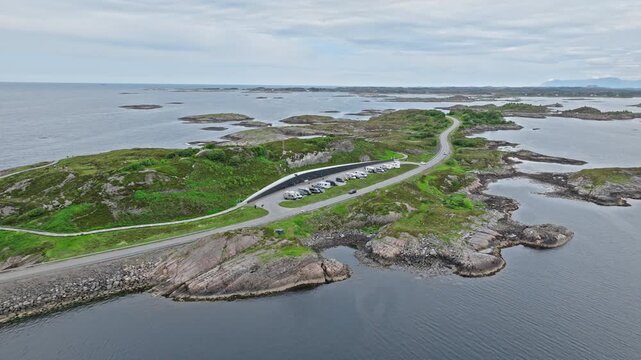 Atlanterhavsveien aerial view of the coastal road threading across green islets with a parking pullout and narrow channels nearby. Drone view rises over the roadway, mapping the bends across skerries.