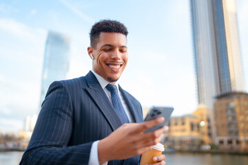 A black man stands near a river in a city. He wears a suit and holds a coffee cup in one hand. He looks at his phone and smiles. Tall buildings are in the background under a clear sky. © Videophilia