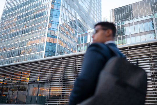 A businessman in a suit walks past modern office buildings during early evening. The glass and steel structures reflect city lights as he carries a bag.