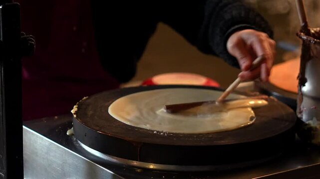 Hands spreading crepe batter on hot griddle with steam and melted chocolate in background