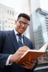 Fototapeta premium A black man in a suit stands outdoors in a city. He smiles while writing in a notebook. Tall buildings surround him. It is daytime and he appears focused on his task.