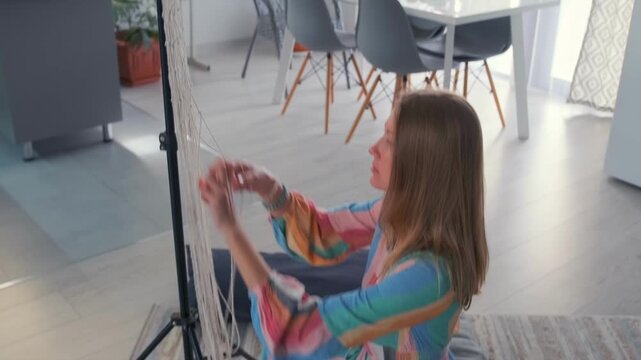 Young woman in colorful dress weaving macrame threads at home. Creative handmade project -  cozy lifestyle and boho interior decoration.
