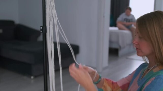 Young woman weaving macrame threads in living space while her friend relaxes in the bedroom in the background. Cozy lifestyle -  handmade craft and friendship.