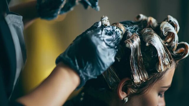 Medium shot of a stylist wearing gloves applying chemical perm solution to hair with timed rods background softly blurred to emphasize the hands and protective gear.