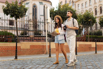 Happy man and woman tourists strolling along cobblestone path in the city, lady holding map, man...