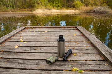 thermos, flashlight and camouflage laser rangefinder lying on a wooden pier on the shore of a lake. outdoor gear placed on a wooden bridge near calm water, natural lakeside environment.