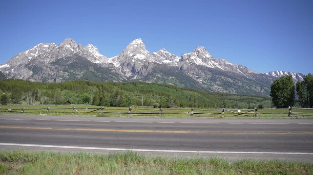 Grand Teton Mountains in the backdrop as a car drives along a scenic road in Grand Teton National Park, Wyoming. Beautiful alpine scenery with a winding road showcasing the iconic mountain landscape.