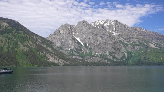 Wide shot of Jenny Lake in Grand Teton National Park, Wyoming, as a tour boat slowly enters the frame. Scenic alpine landscape featuring calm waters, towering mountain peaks, and a peaceful natural at