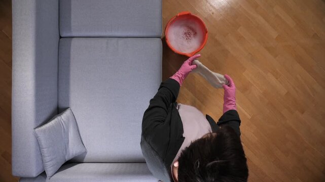 Person wearing pink rubber gloves and black jacket, wringing a damp cloth into an orange bucket filled with soapy water next to a gray couch, maintaining a clean apartment