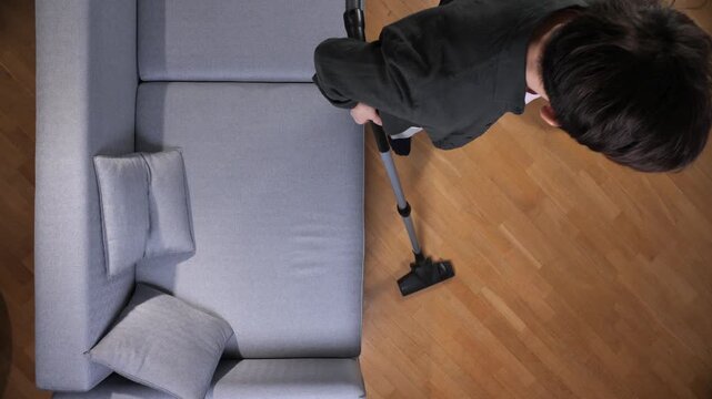 Person cleaning parquet floor in a bright living room, vacuuming dirt and dust from around a comfortable gray couch during routine household chores and home maintenance