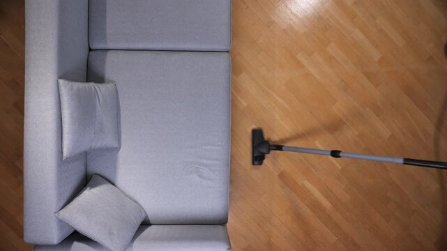 Vacuum cleaner tool running over clean parquet flooring next to a modern gray sectional couch in a bright living room, symbolizing domestic daily chores