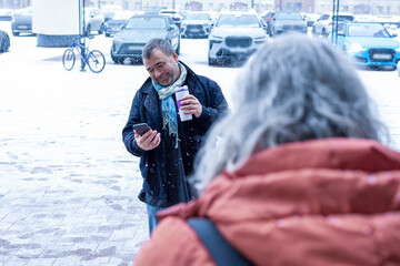 Female photographer in red jacket filming man with phone and coffee cup in snowy weather outside...