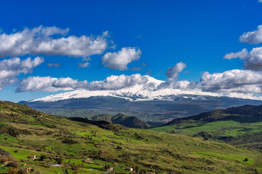 A stunning panoramic view of the active Mount Etna volcano in Sicily, Italy, its peak covered in snow during the winter or spring season. The foreground features vibrant green rolling hills and agricu