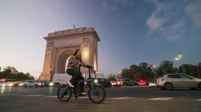 Young woman riding a bicycle on a busy street near the arch of triumph during sunset, surrounded by city traffic
