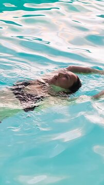 Vertical video of a serene woman in a swimsuit floating on her back in a clear blue swimming pool, relaxing with her eyes closed.